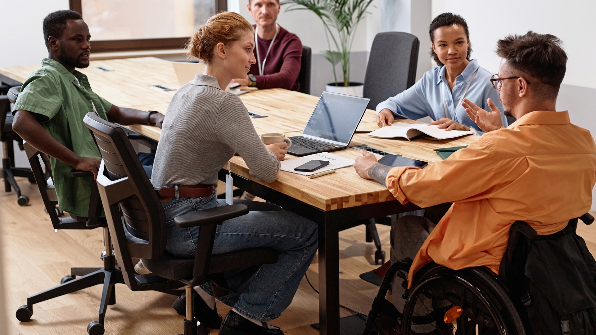 Board meeting comprised of a diverse team of 5 people, including a man using a wheelchair at the head of the table