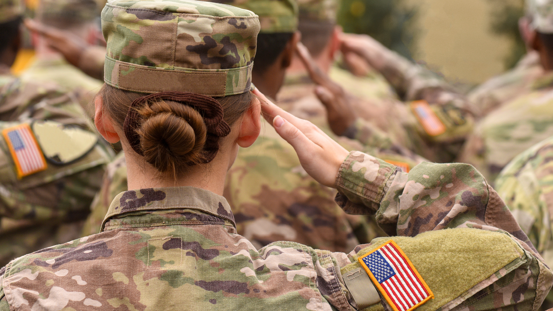 Soldiers saluting the American flag, wearing fatigues, all shown from back with a female being the primary subject in the image